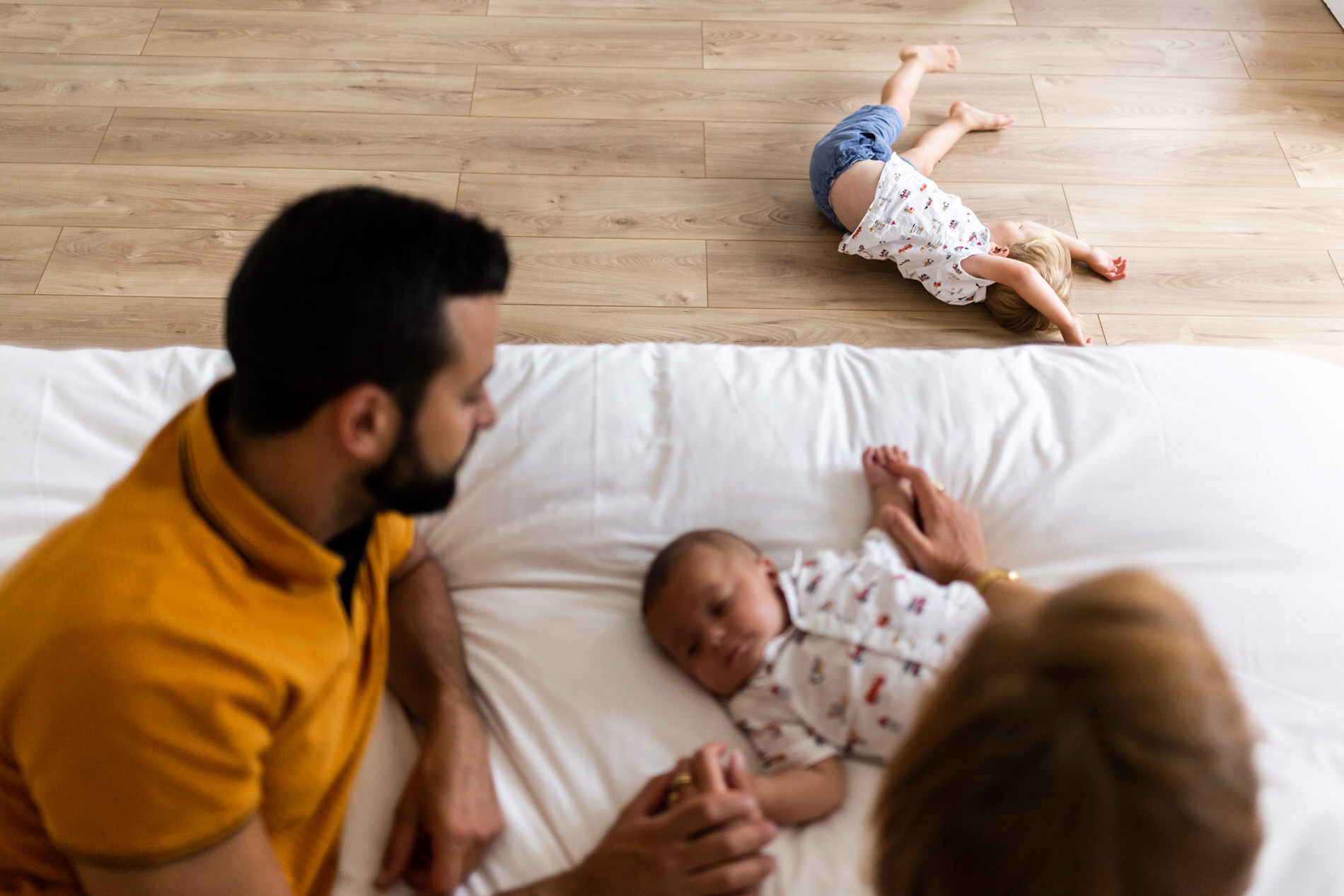 des parents avec bebe sur le lit pendant que le fils aine fait des galipettes par terre pendant une seance photo famille val d oise avec camille piovesan